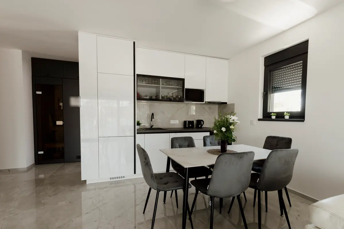 Open plan kitchen with tall white gloss cabinets, a stone-look backsplash, grey chairs at a white table and glossy tile floors