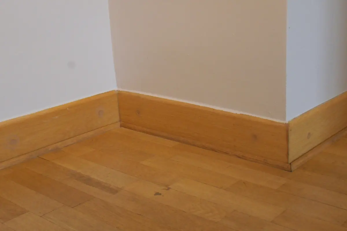 Oak baseboard meeting at an inside corner beneath white and beige walls, beside a parquet floor