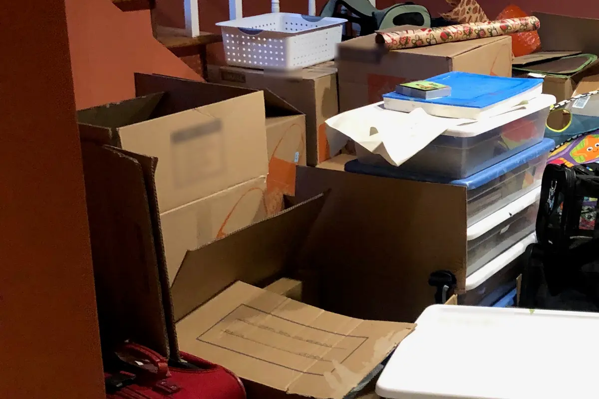 Open and stacked boxes beside basement stairs, with plastic drawers, a white basket and a red suitcase
