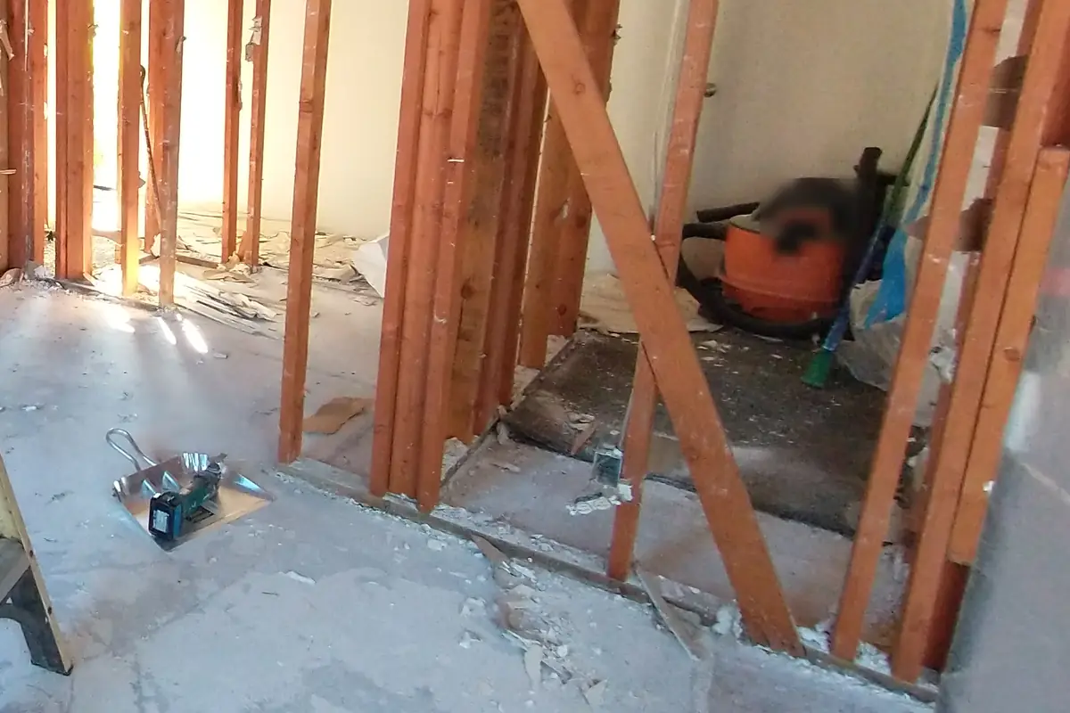Gutted kitchen with exposed studs, drywall scraps and a layer of fine dust across the subfloor beside a wet/dry vacuum