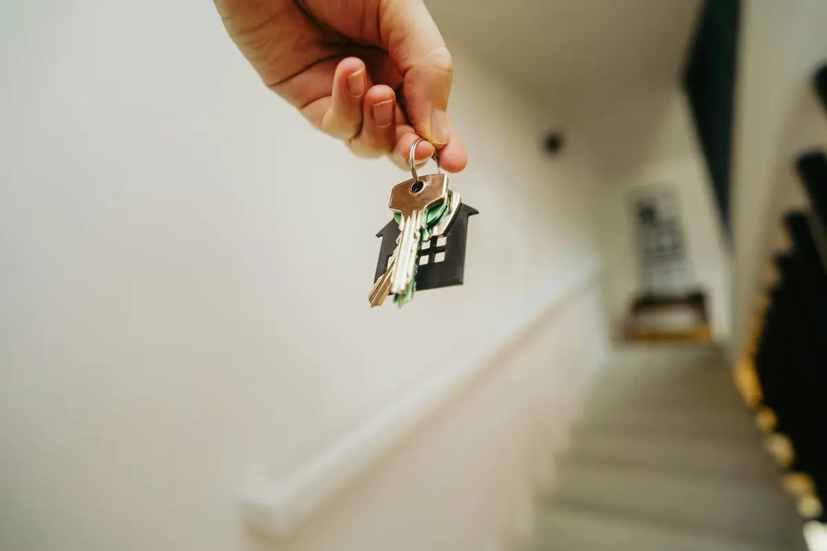 Hand holding keys with a black house-shaped fob against a softly blurred white stairwell and handrail