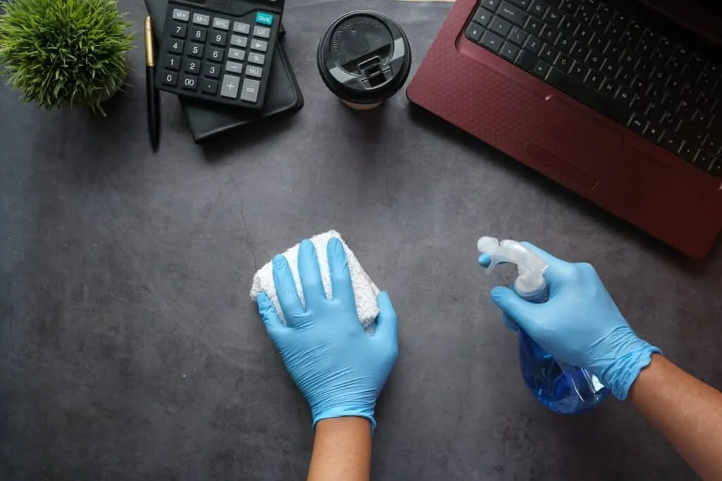 Gloved hands wiping down an office desk next to a laptop