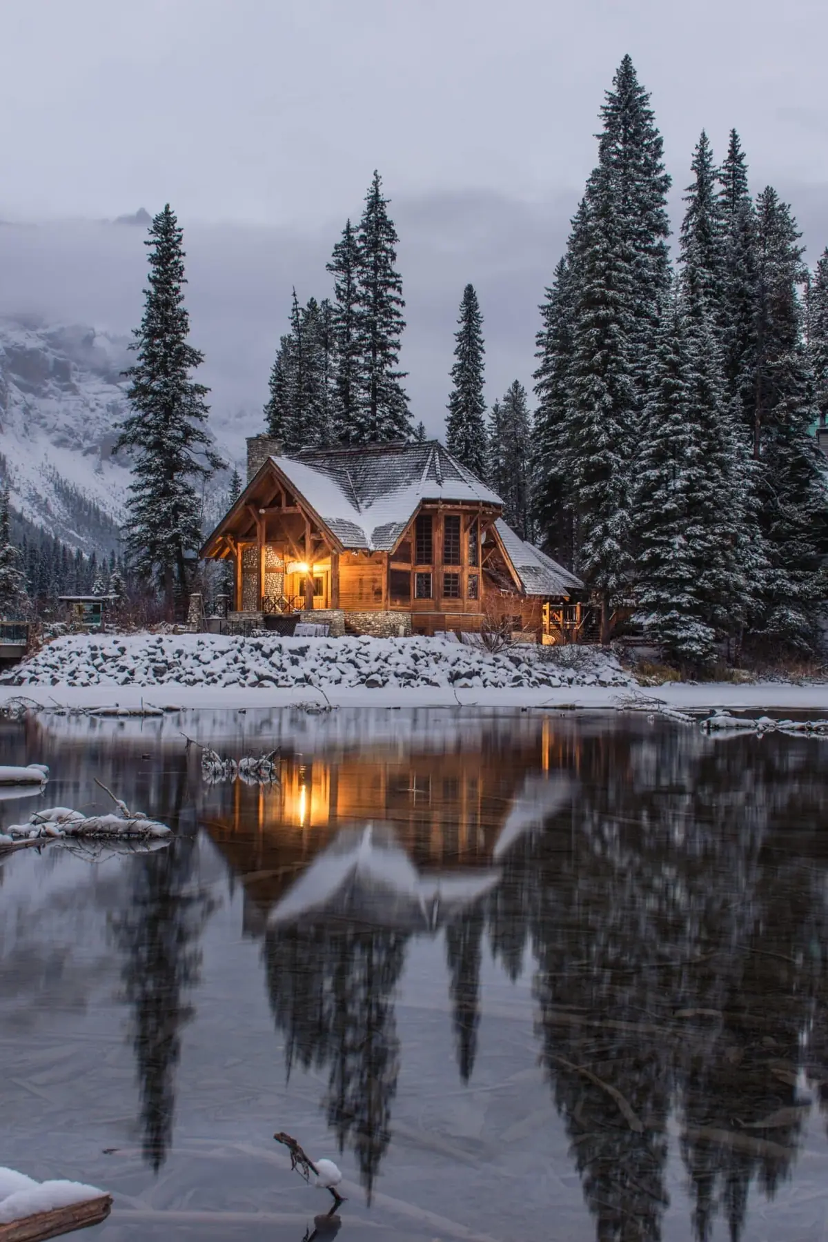 Snow-covered log cabin by a lake surrounded by evergreen trees