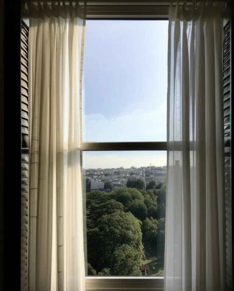 Open window with sheer white curtains overlooking green treetops